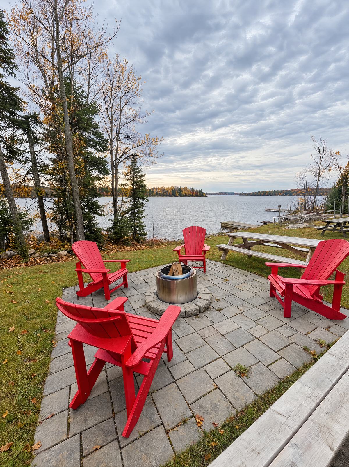 Red Adirondack chairs around a fire pit on a stone patio by the lake, with fall colour on the far shore