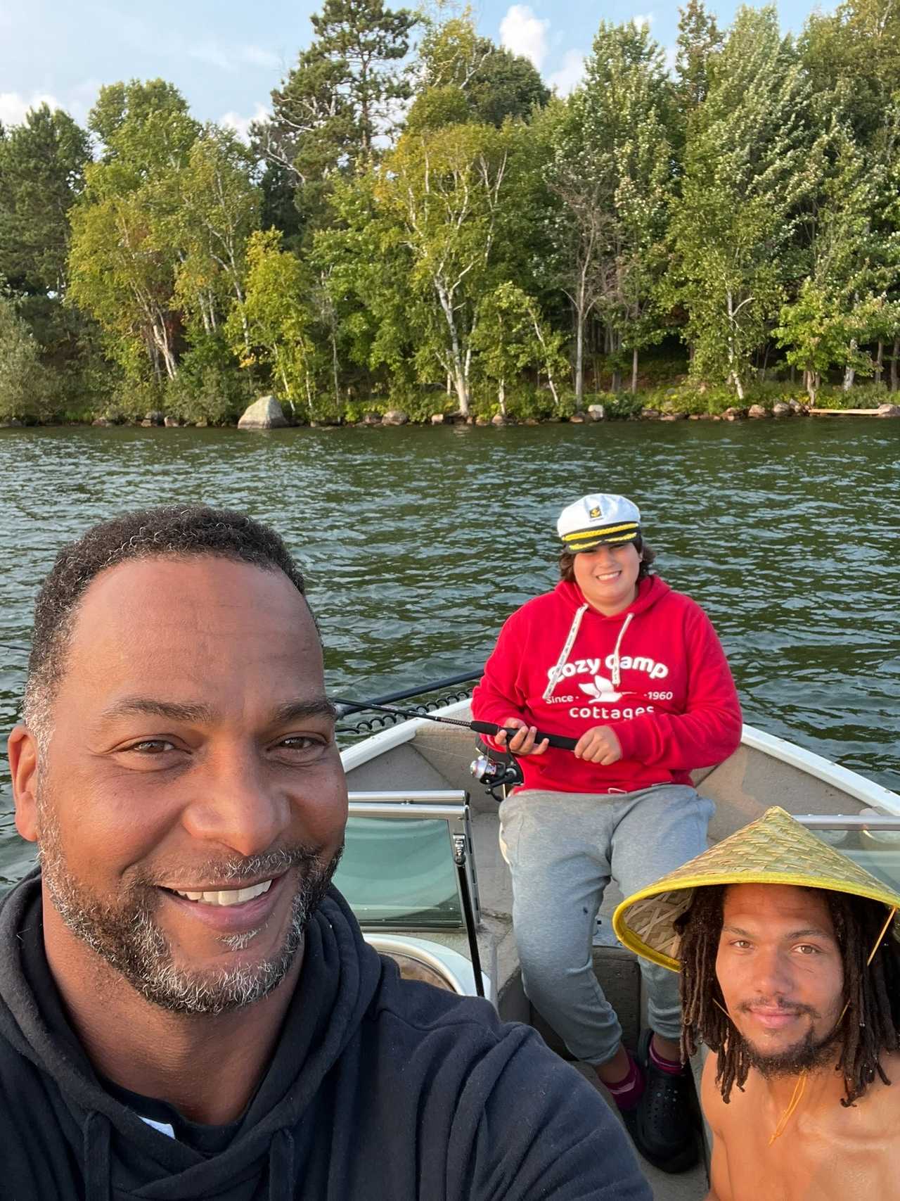 Three smiling people taking a photograph together in a fishing boat by a wooded shoreline