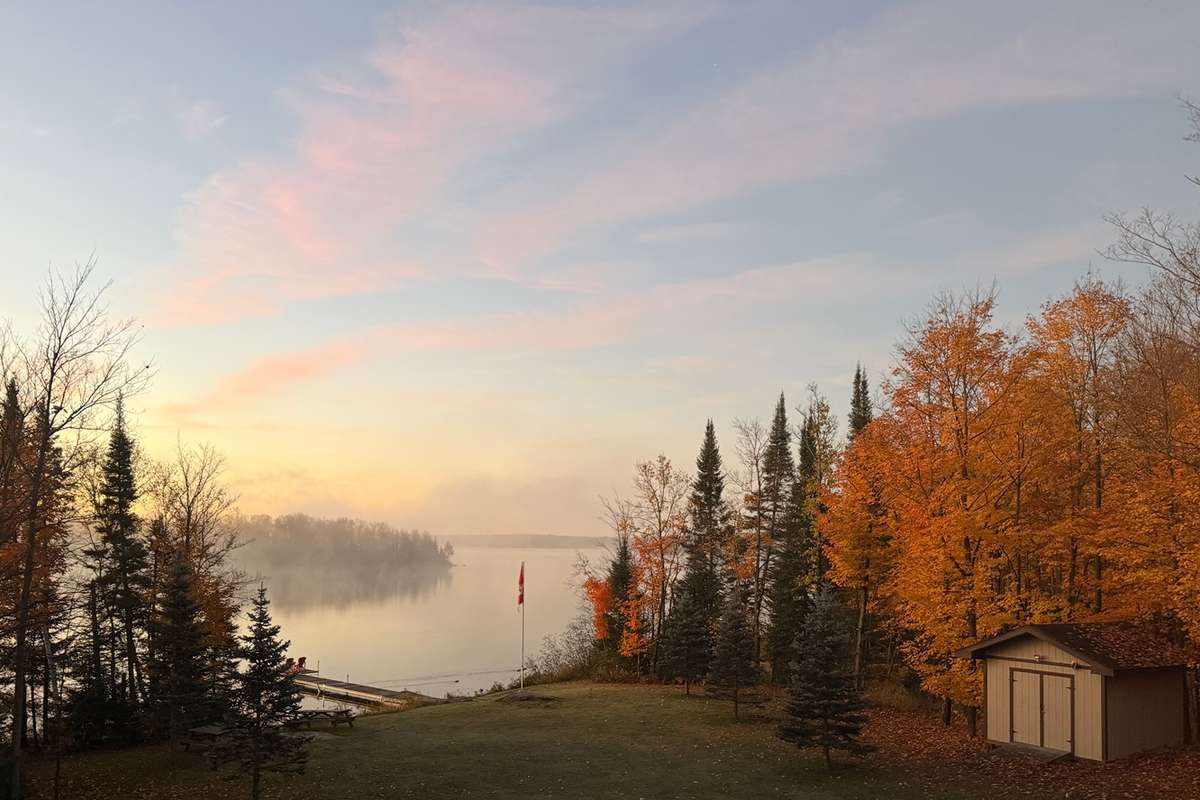 Mist over a calm lake and a wooded island at dawn, seen down a lawn to a dock and a flagpole, between evergreens and orange autumn maples under a pink-streaked sky