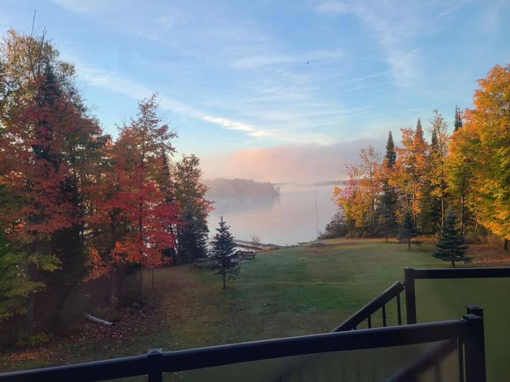 Red, orange and yellow autumn trees framing a misty lake and a wooded island at dawn, seen from a deck above a lawn sloping down to the docks