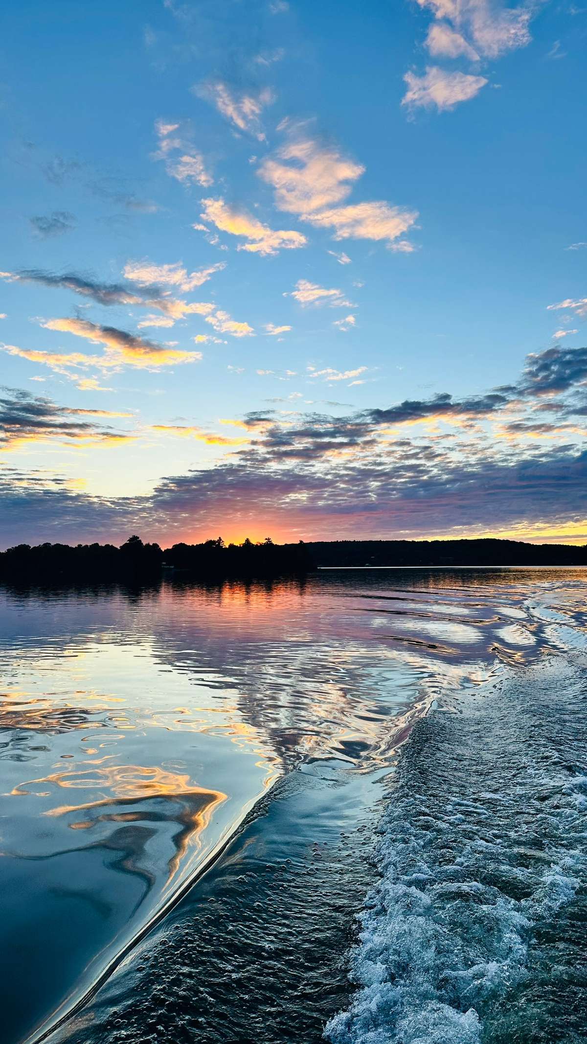 A boat's wake curving across calm water at sunset, the clouds mirrored in pink, gold and blue, with a dark wooded shoreline ahead