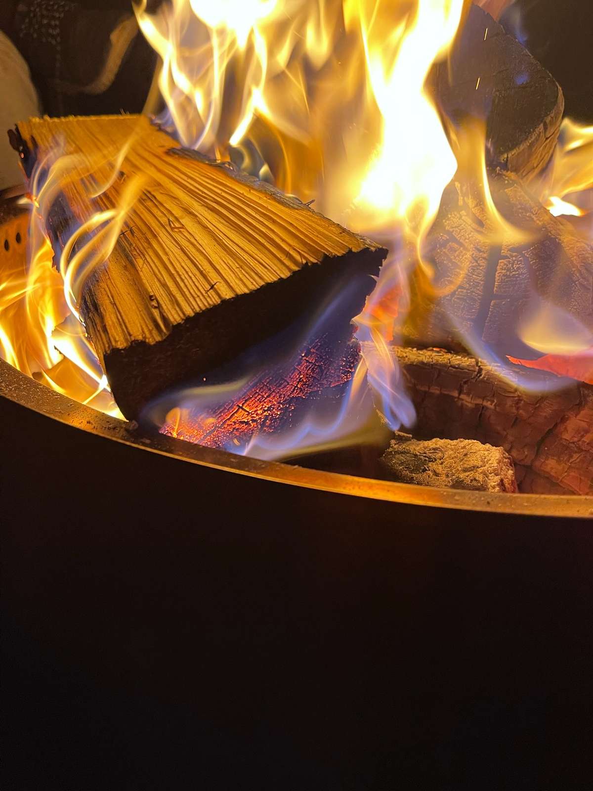 Split logs burning in a steel fire bowl, tall yellow flames rising around them and embers glowing red beneath