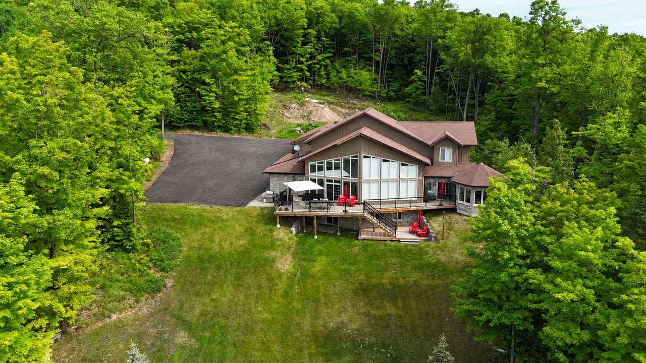 Clubhouse from the air: the house with its wall of tall windows, decks and red chairs at the top of a long lawn, with woods all around