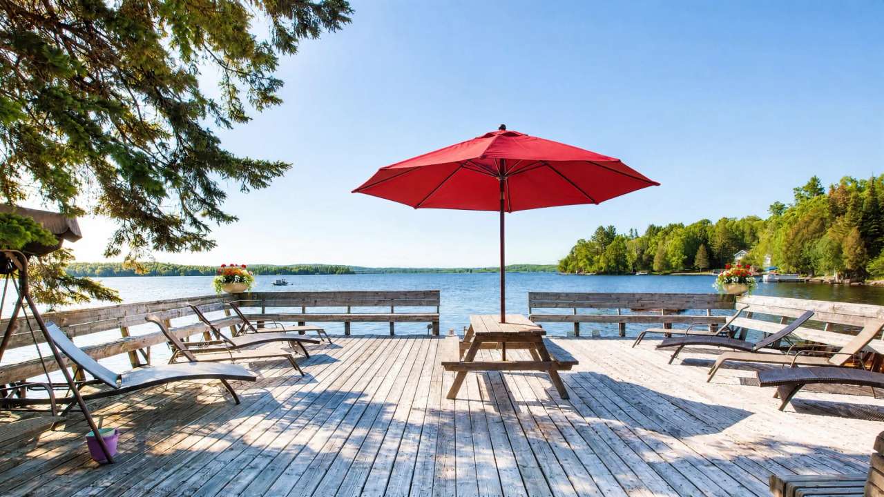 The lakefront sun deck with a red umbrella open over the picnic table, flowers in the railing planters and lounge chairs either side, Lake Nosbonsing beyond