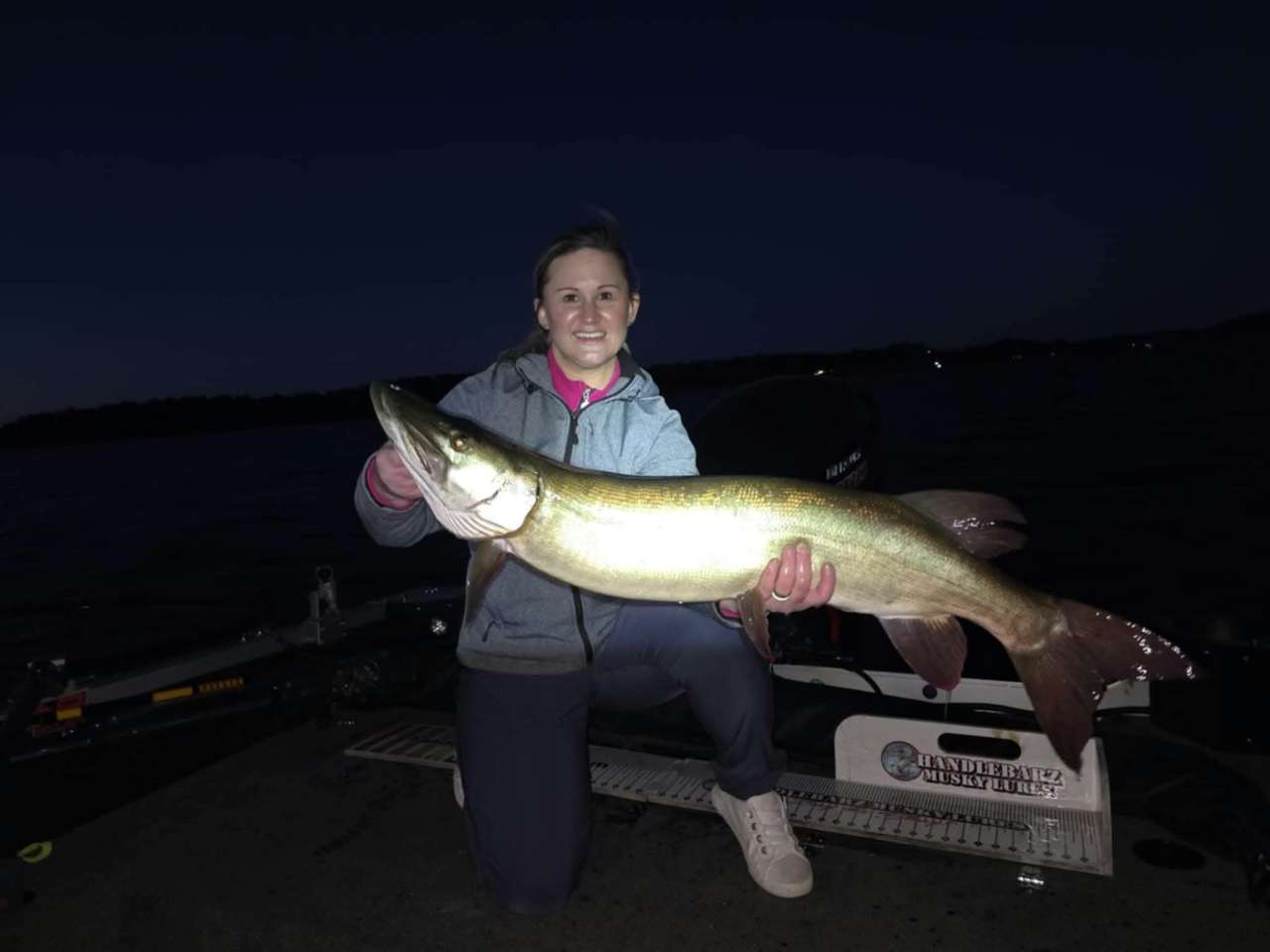 A smiling angler kneeling on a boat deck at dusk, holding a long musky across both hands, with the dark lake and shoreline behind