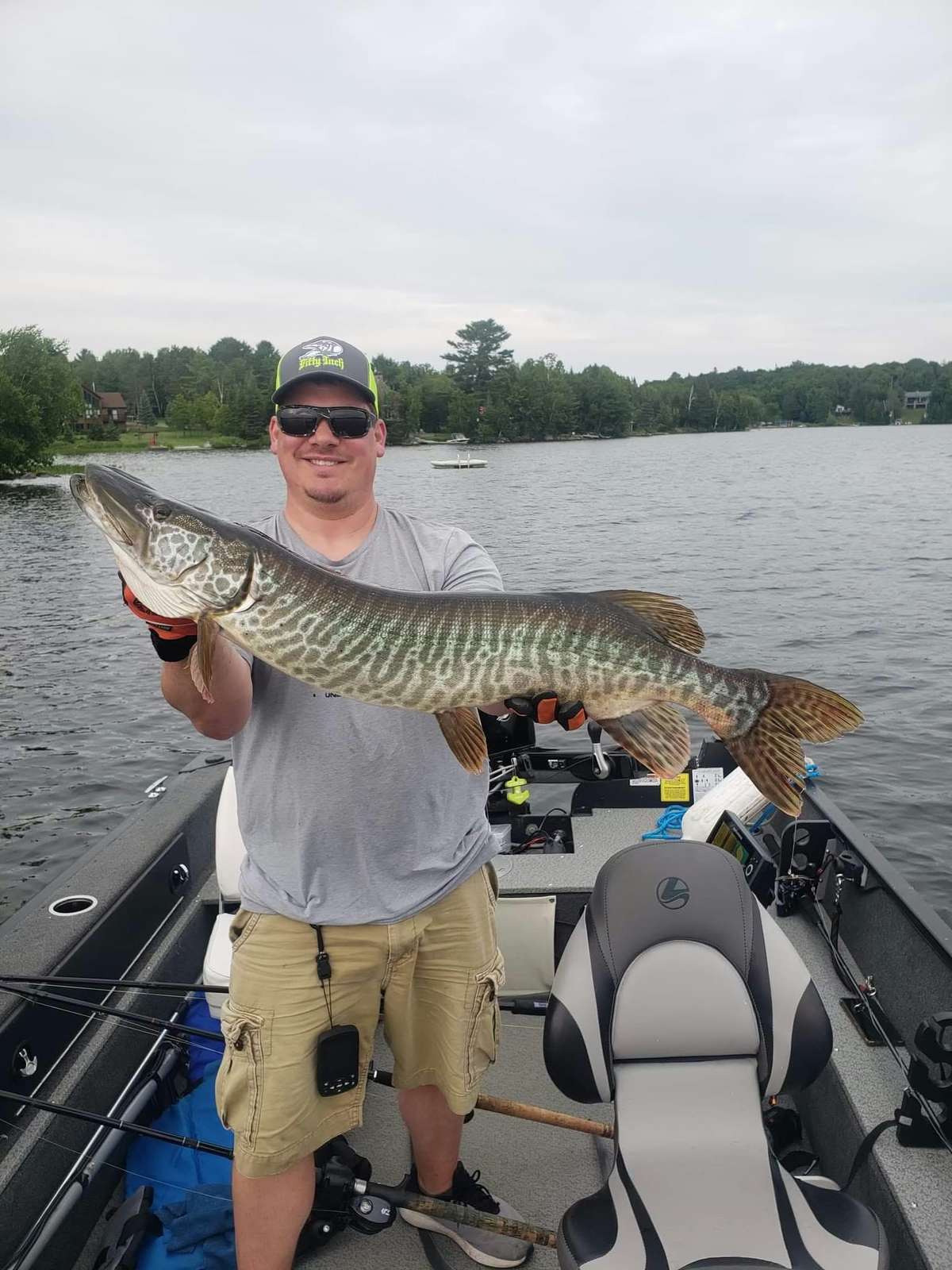 A smiling angler in a cap, sunglasses and grey T-shirt standing in a fishing boat, holding up a long, boldly barred musky in gloved hands, with the lake and a wooded shore with cottages behind
