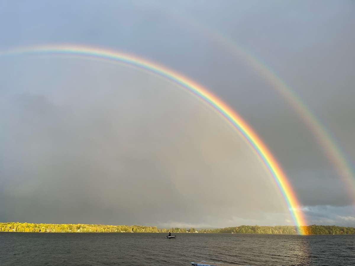 A bright double rainbow arching over the lake under grey storm clouds, with a small boat on the water and a sunlit wooded shore beyond