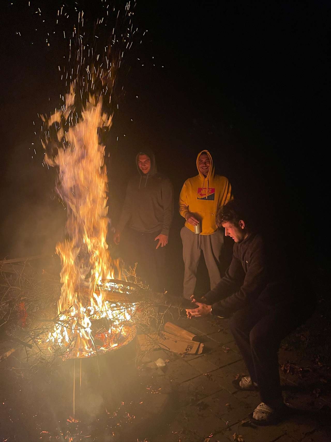 Three friends in hooded sweatshirts laughing beside a tall campfire at night, one crouching with hands held out to the flames as sparks rise