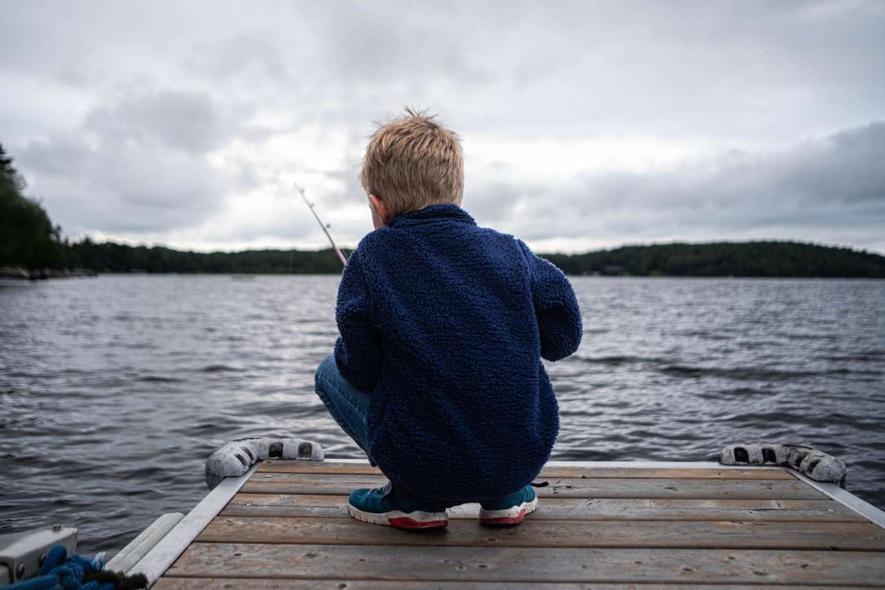 A child in a navy fleece crouching at the end of a dock with a fishing rod, looking out over the lake under grey clouds