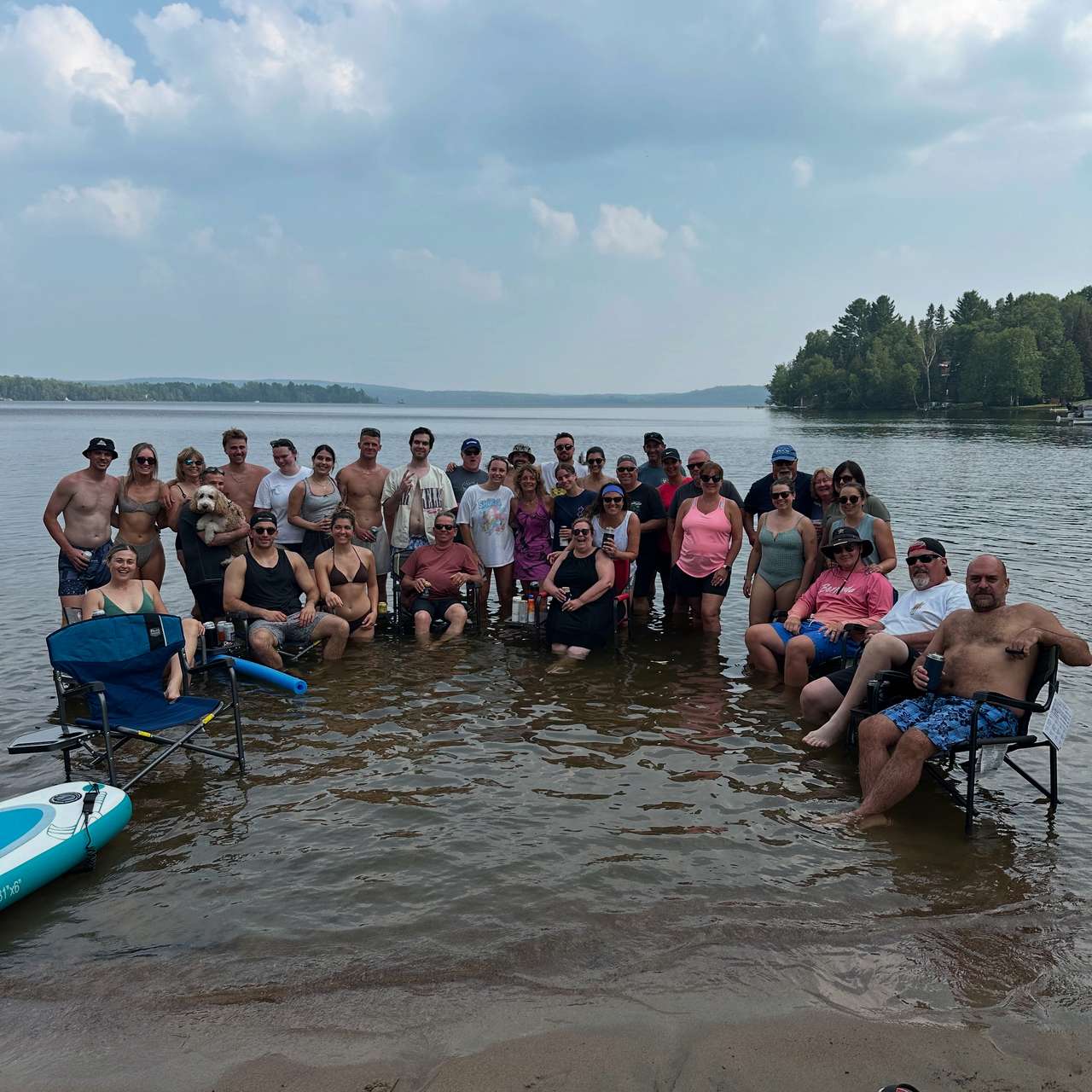 A large group of guests standing and sitting in camp chairs in the shallow water off a sandy beach, smiling, with the lake and a wooded point behind