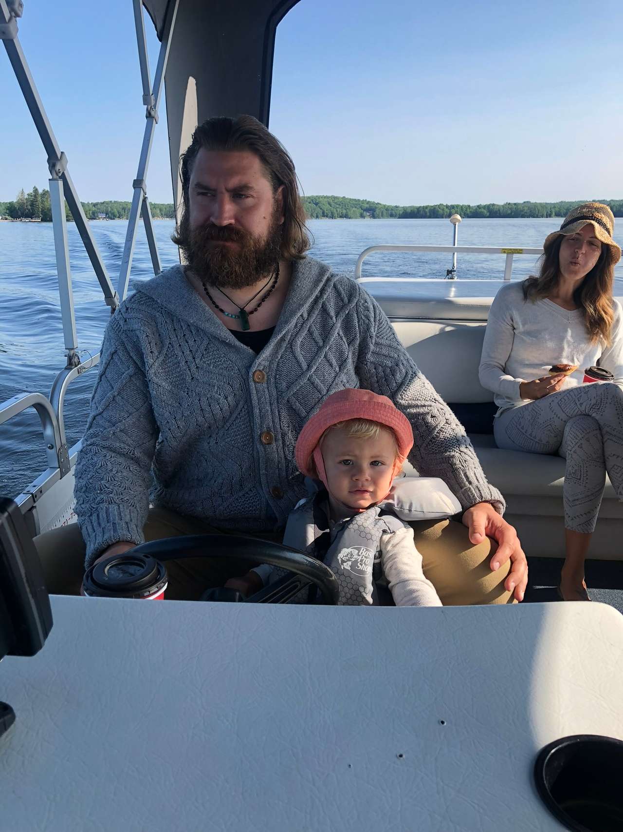 A toddler in a pink sun hat and life jacket at a pontoon boat's wheel on a bearded adult's lap, with another passenger in a straw hat behind them and the lake beyond