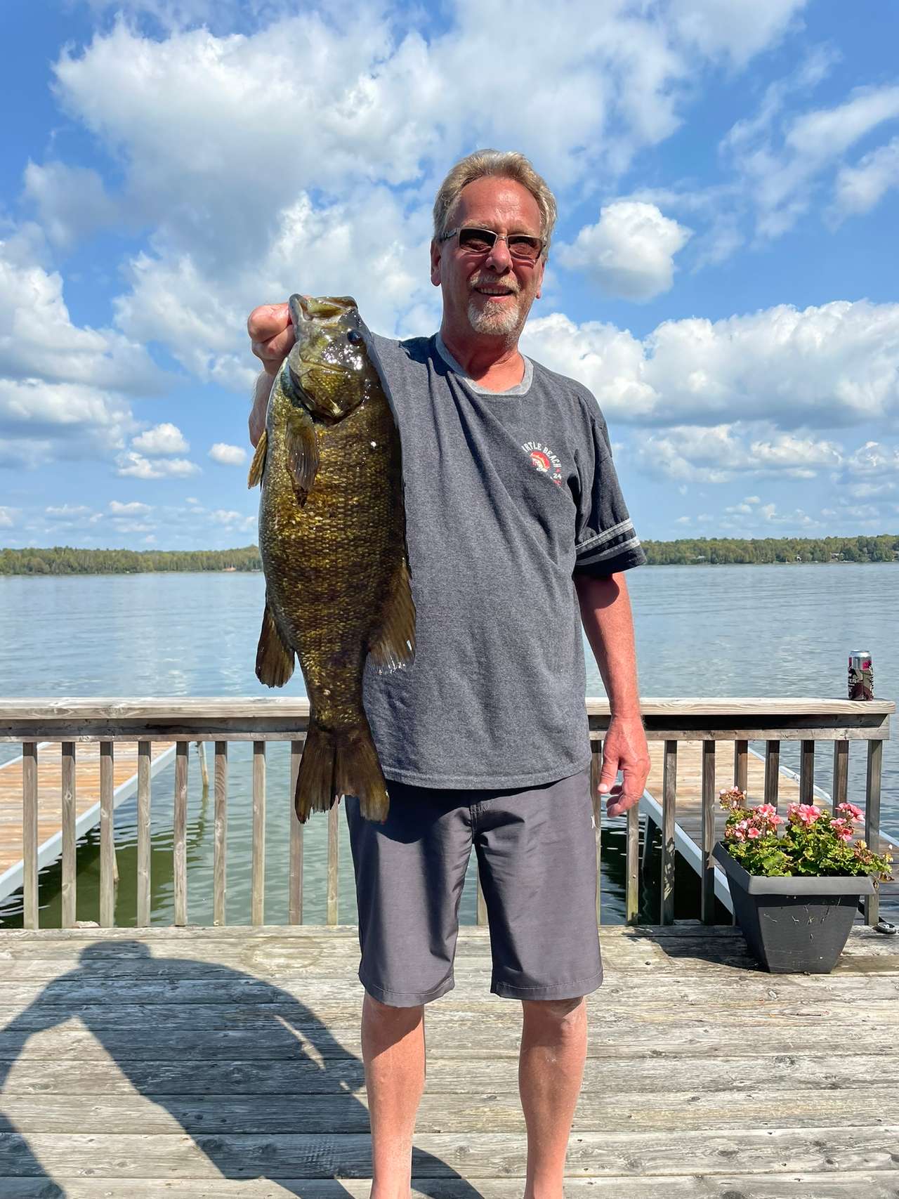 A smiling angler in sunglasses holding up a large fish on a lakeside dock under a blue sky with white clouds