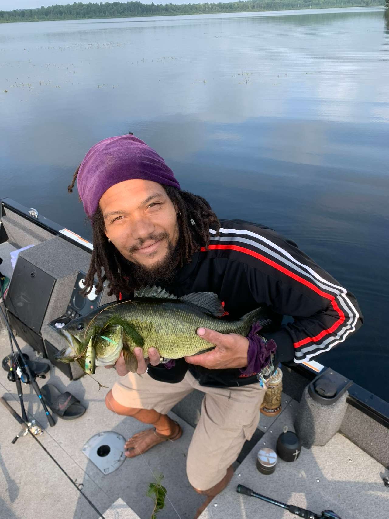 A smiling angler in a purple headscarf crouching on a boat deck with a fish held in both hands, calm lake water behind