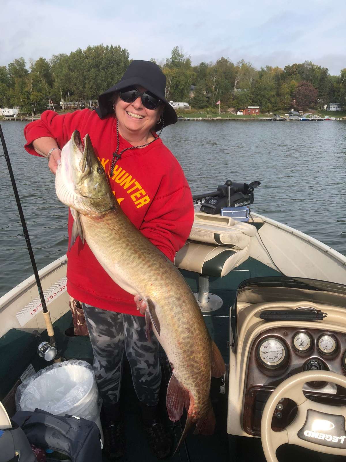 A smiling angler in a red sweatshirt, wide-brimmed hat and sunglasses holding up a long fish in a boat, with the lake and the far shore behind