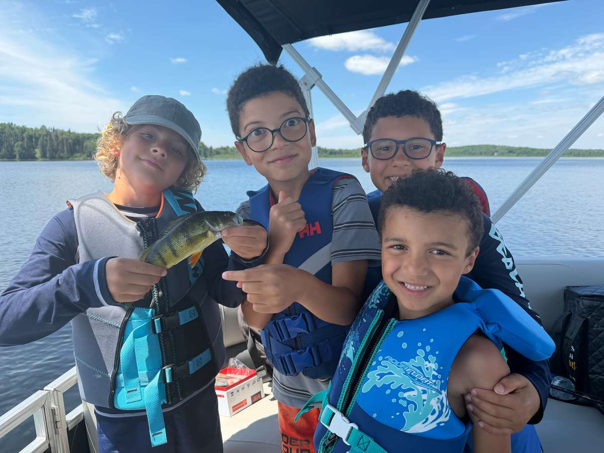 Four children in life jackets on a boat under a canopy, one holding up a small fish, with the lake and a wooded shore behind them