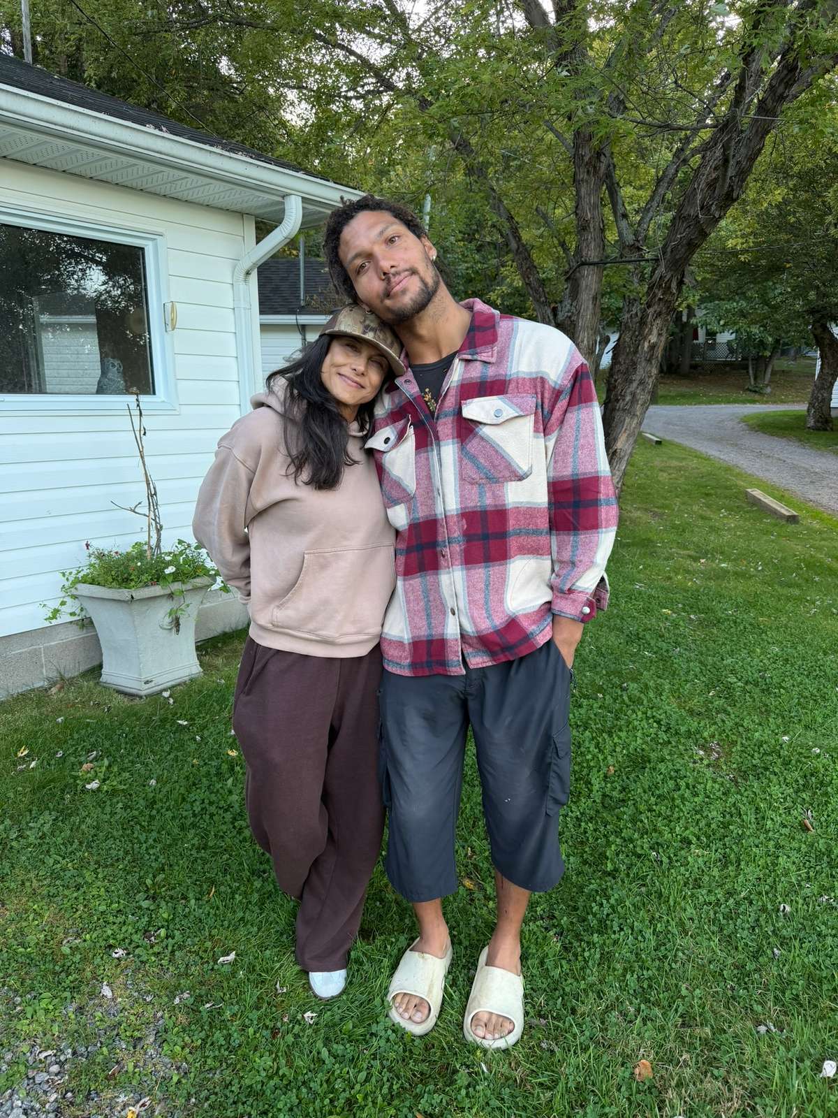 Two guests leaning their heads together on the lawn beside a white cottage, one in a camouflage cap and hoodie, the other in a red plaid shirt, under shady trees