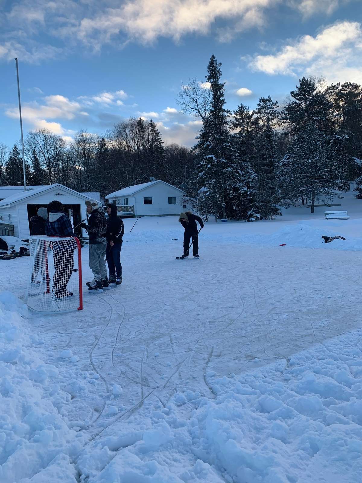 Friends in winter coats and skates playing hockey on an outdoor rink cleared in the snow, beside a net, white cottages and tall snow-covered pines