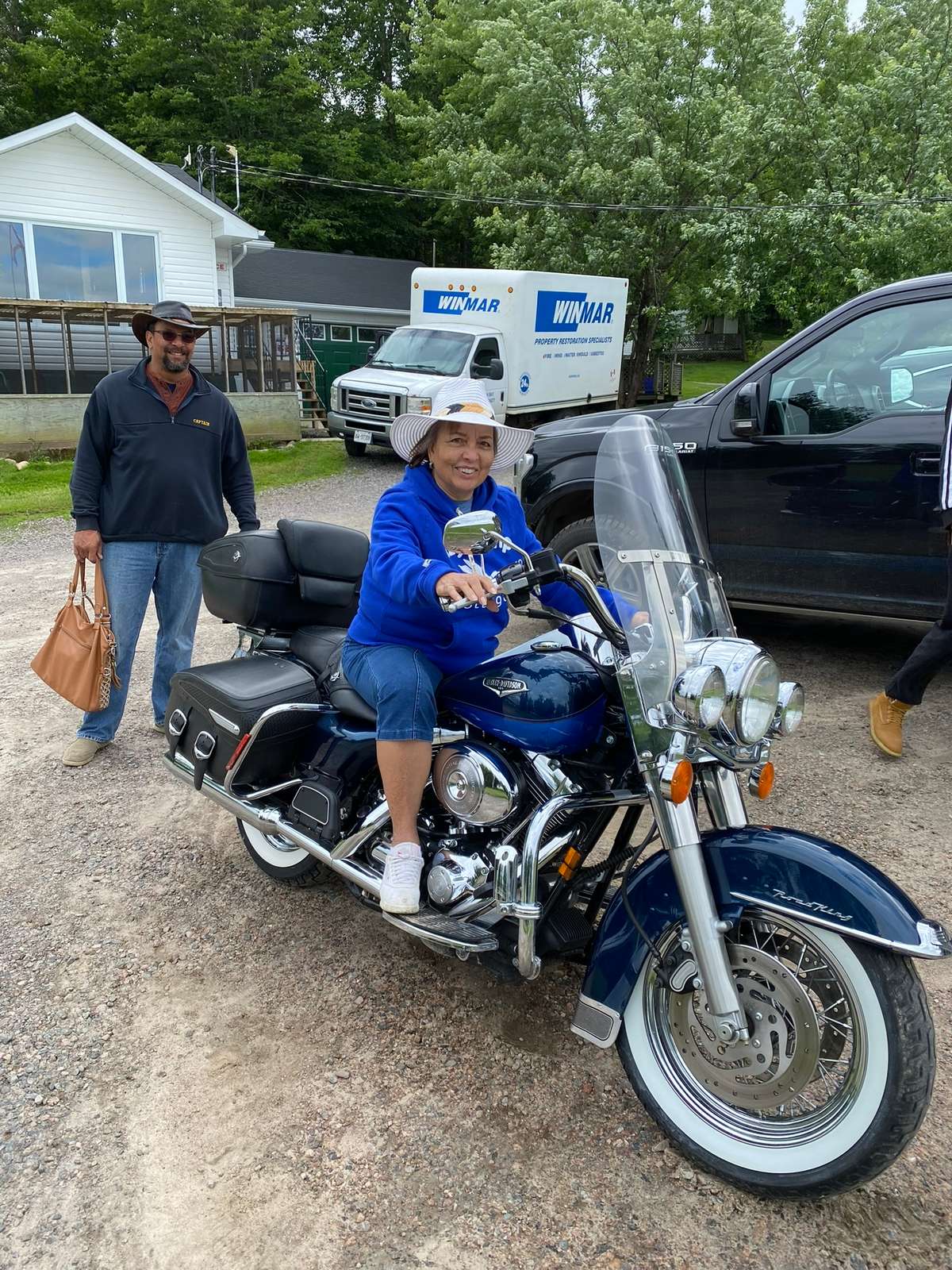 A smiling guest in a wide-brimmed sun hat and blue hoodie sitting on a navy touring motorcycle on a gravel lot, with a friend in a brown hat standing behind and a white cottage beyond