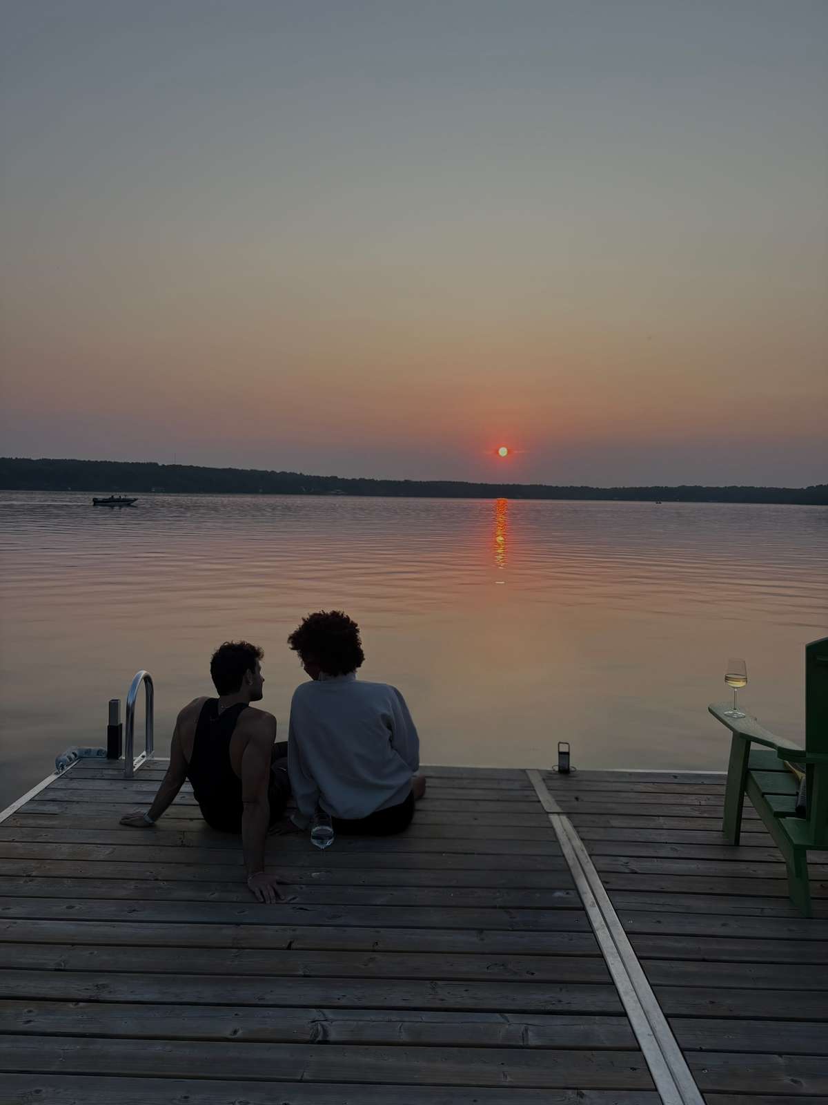 Two guests sitting side by side at the end of a dock watching the sun set over a calm lake, with a glass of wine on the arm of a green Adirondack chair beside them