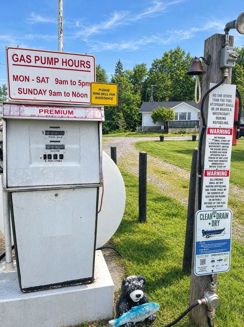 The camp's fuel pump under its hours sign, beside a wooden post with a bell, a fuel hose and refuelling safety signs, a carved bear holding a Welcome fish at its foot and a white house across the lawn