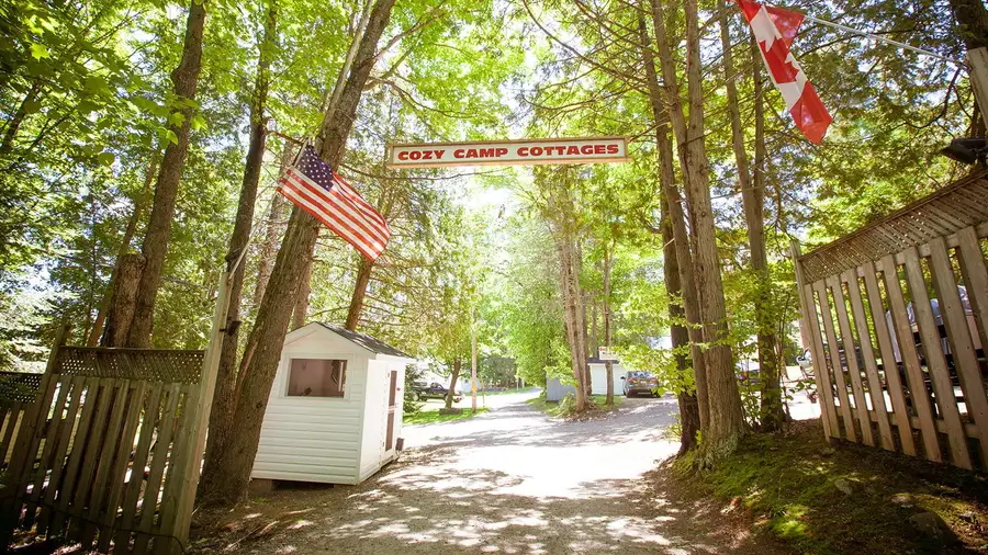 The Cozy Camp Cottages entrance sign over the tree-lined lane, with Canadian and American flags