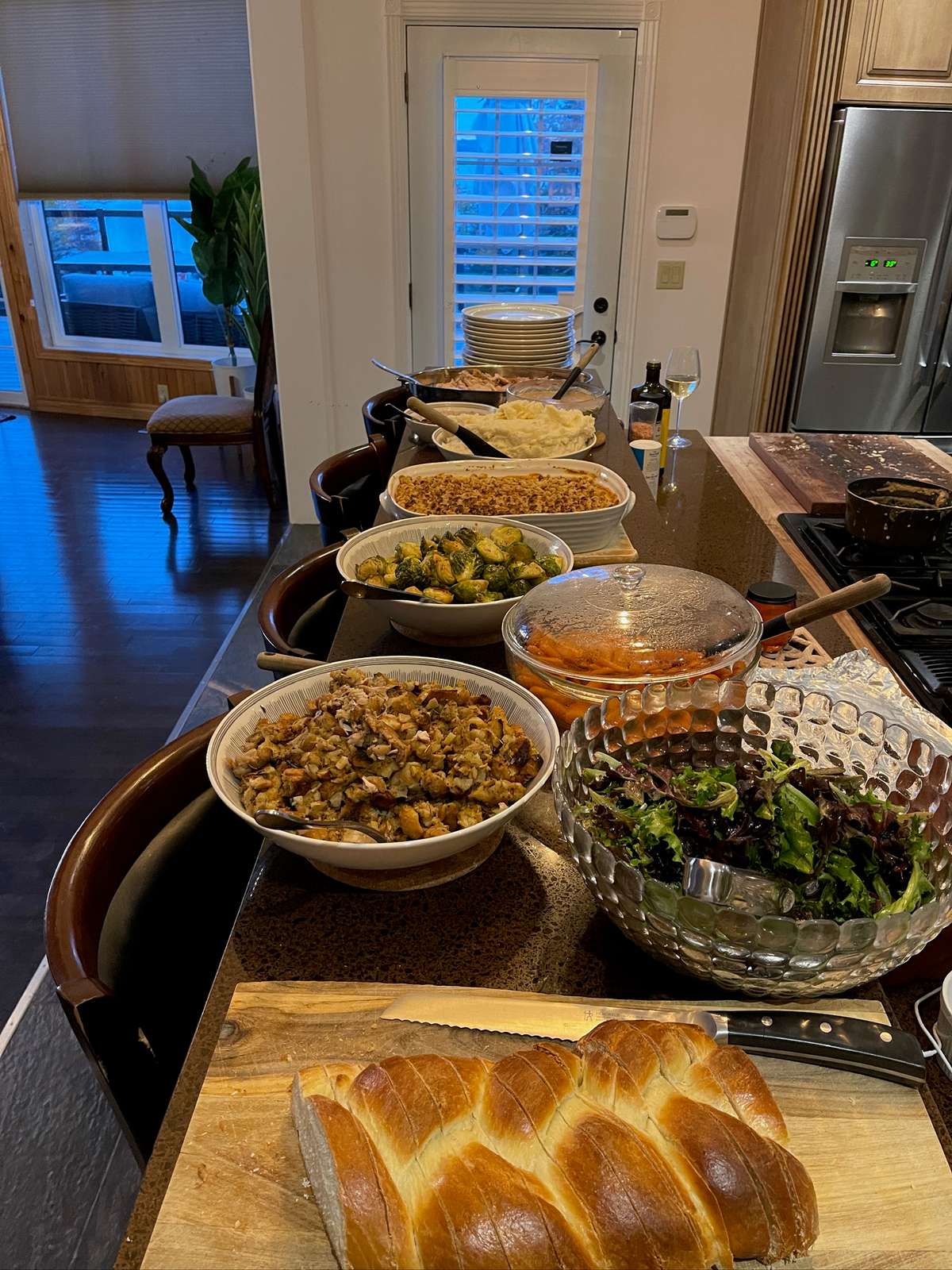 A dinner for a crowd laid out along a kitchen island: mashed potatoes, a baked casserole, Brussels sprouts, carrots, stuffing, a green salad and a braided loaf