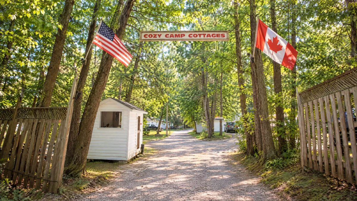 The Cozy Camp Cottages sign over the gravel lane at the camp entrance, between American and Canadian flags, in morning sun under tall trees