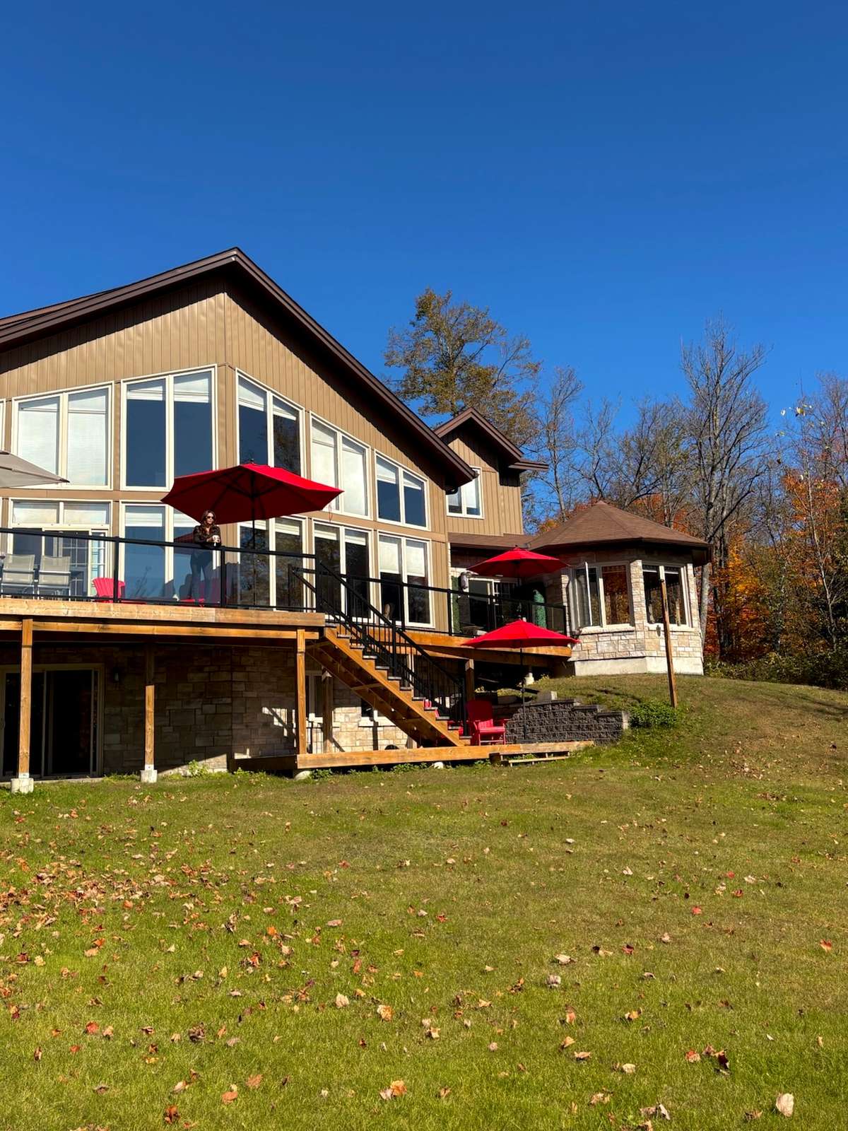 The lakeside back of a large lake house with tall windows, a raised deck with red umbrellas and a guest at the railing, stairs down to a lower patio and a stone gazebo, under a clear blue sky
