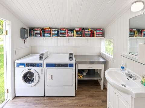 Inside the guest laundry hut: a washer and dryer side by side, a steel folding table, a sink and a long shelf of books under the white sloped ceiling