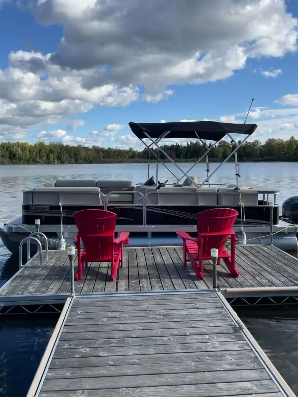 A pontoon moored beyond the dock and red chairs on Lake Nosbonsing