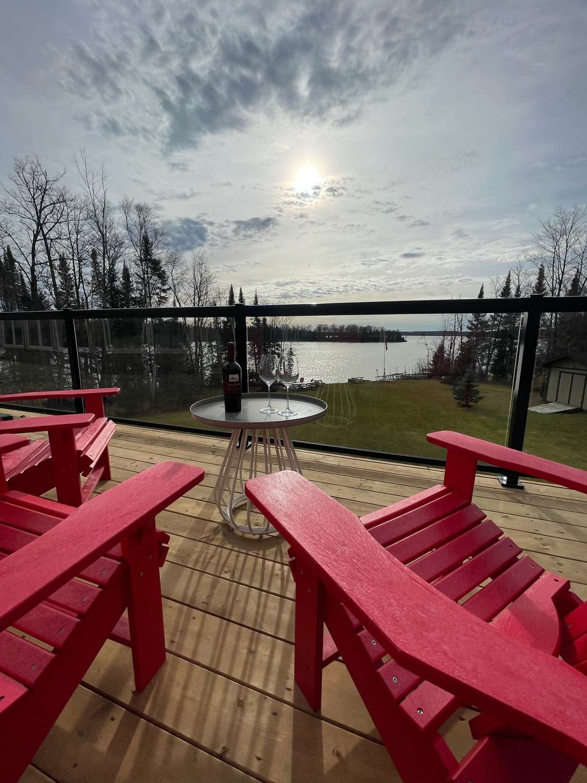 Red Adirondack chairs around a small round table set with a bottle of red wine and two glasses on a deck, a glass railing and a lawn sloping down to the lake beyond
