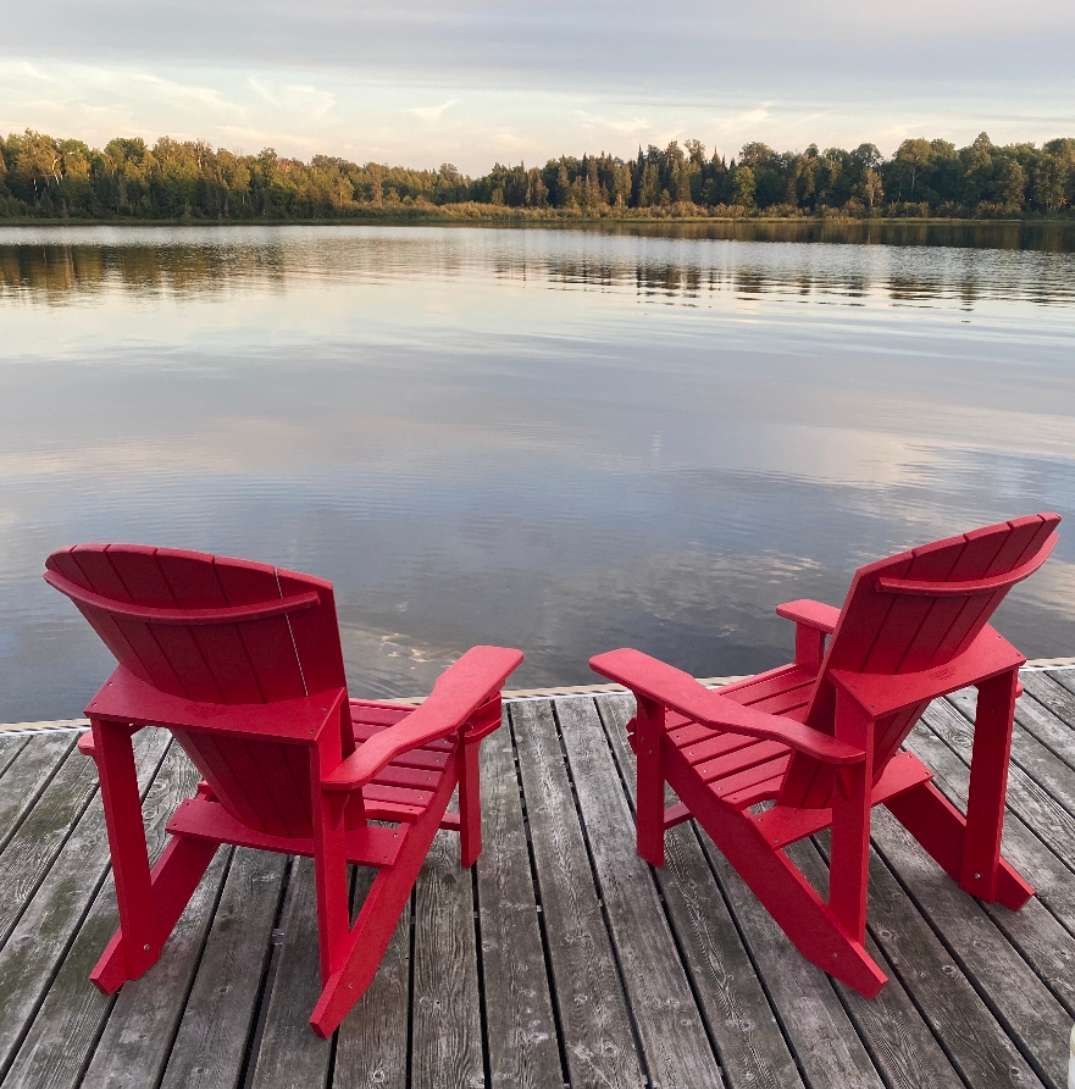 Two red Adirondack chairs on a wooden dock, facing a calm lake