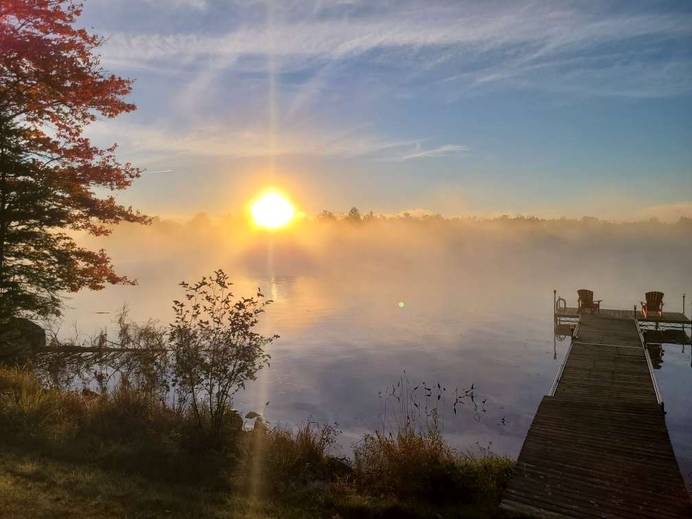 The sun rising through mist over a calm lake, with two Adirondack chairs at the end of a wooden dock and red autumn leaves overhead