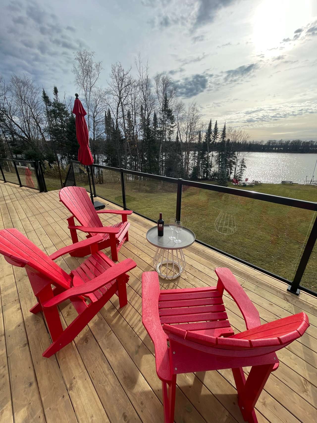 Three red Adirondack chairs around a small round table with a bottle of red wine and two glasses on a wooden deck, a glass railing and a closed red umbrella beyond, and the lake through the trees